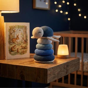 Op de afbeelding: Een handgemaakt stapelspeelgoed van haakwerk in blauw- en crèmetinten, dat op een schildpad lijkt. Het speelgoed staat op een houten oppervlak naast een kinderboek en een kleine lamp. De kamer heeft een kinderkamerthema.