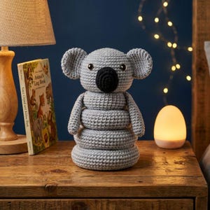May include: A gray crocheted koala bear with a black nose and button eyes. The koala is sitting on a wooden table next to a lamp and a children's book. The koala is a handmade item.