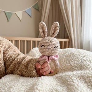 Può includere: Un sonaglio a forma di coniglietto all'uncinetto beige con un colletto rosa a balze e un anello di legno. Il coniglietto ha occhi ricamati e un viso sorridente. Il sonaglio è tenuto in mano da un bambino su una coperta bianca e soffice.