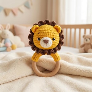 Peut inclure: Un hochet en forme de lion crocheté en jaune et marron, avec un anneau en bois. Le lion a un visage détaillé avec un museau blanc et des yeux foncés. Le hochet est conçu pour les bébés et est posé sur une couverture crème. L'anneau en bois mesure environ 7 cm de diamètre.