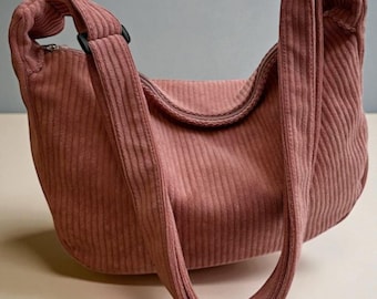 May include: A dusky pink corduroy shoulder bag with a curved design. The bag has a long strap and a zip closure. The corduroy fabric has vertical ridges, and the strap is attached with black fittings.