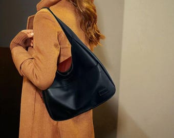 Pode incluir: Uma bolsa de ombro preta de couro com um design curvo. A bolsa tem uma única alça e um bolso frontal. Usada sobre um casaco marrom, o interior da bolsa é vermelho. Um acessório elegante.