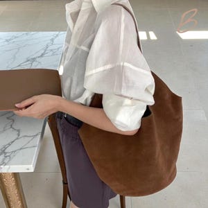 Peut inclure: Un grand sac fourre-tout en daim marron avec une bandouli&egrave;re. Le sac est d'un brun chaud et riche et semble vide. La personne sur l'image porte un chemisier blanc et un short fonc&eacute;.