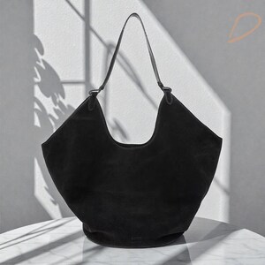 Peut inclure: Un sac &agrave; bandouli&egrave;re en daim noir avec un design incurv&eacute;. Le sac a une seule sangle et un int&eacute;rieur spacieux. Le sac est pos&eacute; sur une surface blanche avec des ombres.