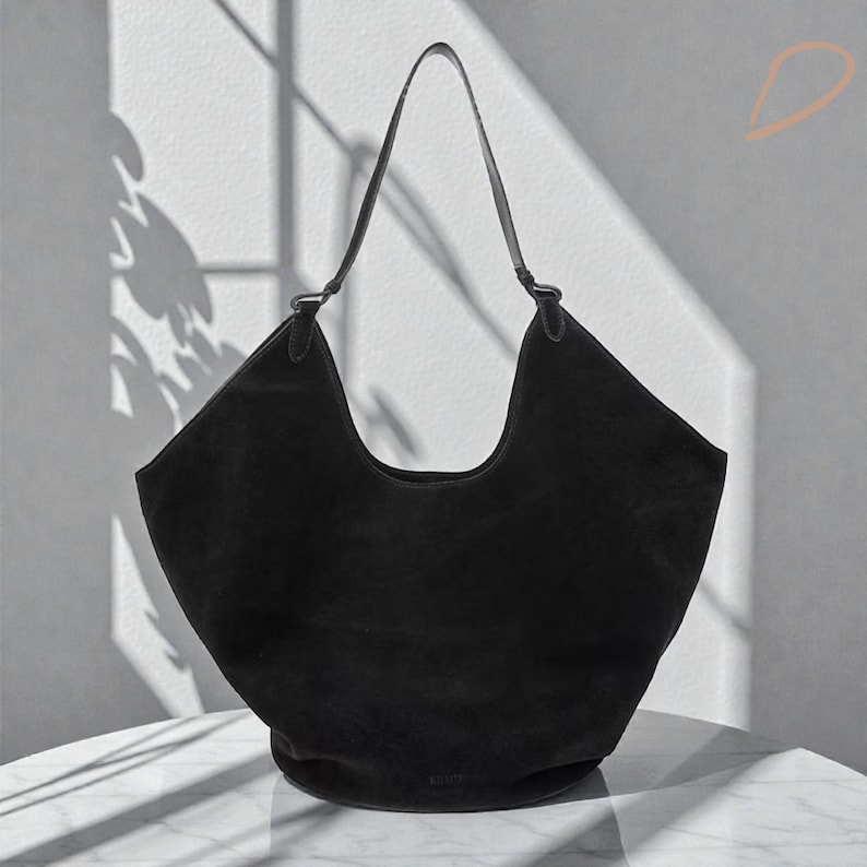 Peut inclure: Un sac &agrave; bandouli&egrave;re en daim noir avec un design incurv&eacute;. Le sac a une seule sangle et un int&eacute;rieur spacieux. Le sac est pos&eacute; sur une surface blanche avec des ombres.
