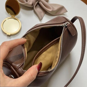 May include: A brown cylindrical handbag with a zipper closure, open to reveal a tan interior. A small, gold compact mirror and a pink hair accessory are in the background. The bag has a small handle.