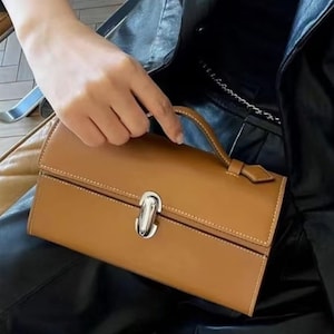 Peut inclure: Un sac à main en cuir marron clair avec un fermoir argenté et une poignée supérieure. Le sac rectangulaire a des coutures blanches sur les bords et une fermeture à rabat. Le sac est tenu par une personne.