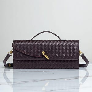 May include: A dark burgundy woven leather handbag with a top handle and a detachable shoulder strap. The bag features a gold-toned clasp and hardware. The rectangular shape and intricate weave pattern give it a classic, elegant look.