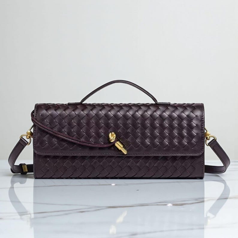 May include: A dark burgundy woven leather handbag with a top handle and a detachable shoulder strap. The bag features a gold-toned clasp and hardware. The rectangular shape and intricate weave pattern give it a classic, elegant look.