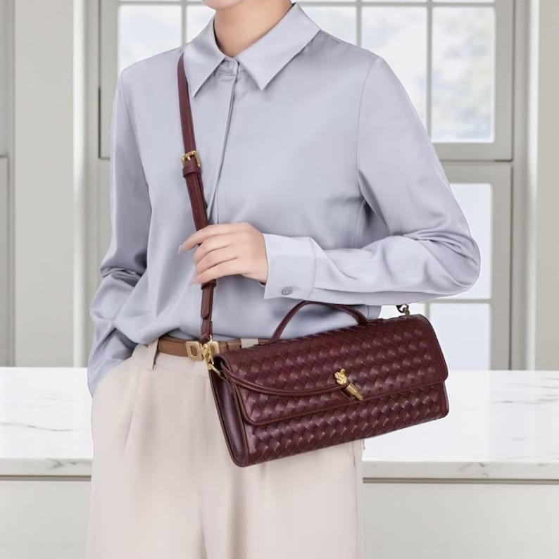 May include: A burgundy leather crossbody bag with a woven pattern. The bag features a top handle and an adjustable strap. It is secured with a gold-coloured clasp. The person is wearing a light blue shirt and beige trousers.