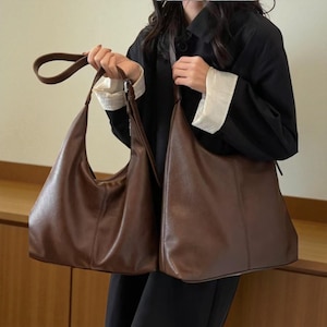 May include: Two brown leather shoulder bags. One bag has a curved handle and a crescent shape. The other bag is a larger, rectangular tote bag. Both bags are a rich, warm brown color and appear to be made of a smooth leather material.