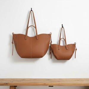 Peut inclure: Deux sacs fourre-tout en cuir marron de différentes tailles suspendus contre un mur blanc. Les sacs ont de longues poignées et des liens latéraux décoratifs. Le plus grand sac mesure environ 46 cm de large, et le plus petit environ 30 cm.