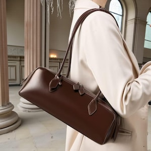 Può includere: Una borsa in pelle marrone scuro di forma rettangolare con una lunga tracolla. La borsa ha una chiusura a cerniera e cuciture decorative. La borsa viene portata a spalla.