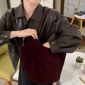 Peut inclure: Un sac fourre-tout en daim bordeaux avec une seule bandouli&egrave;re. Le sac est ouvert, r&eacute;v&eacute;lant l'int&eacute;rieur. La personne porte une veste en cuir marron fonc&eacute;.