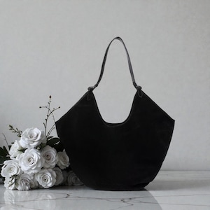Peut inclure: Un sac fourre-tout en daim noir avec un dessus incurv&eacute; et une seule poign&eacute;e. Le sac est pos&eacute; sur un fond neutre avec un bouquet de roses blanches &agrave; gauche. Le design du sac est simple et &eacute;l&eacute;gant.