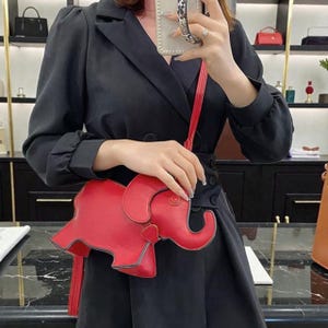 May include: A vibrant red elephant-shaped crossbody bag with a long strap. The bag is crafted from smooth leather and features detailed stitching. The elephant design includes a trunk, ears, and a tail. The bag is held by a person wearing a black blazer.