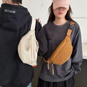 May include: Two corduroy fanny packs in cream and tan. The bags have a zippered main compartment and an adjustable strap with a buckle. The cream bag is worn over a black hoodie, and the tan bag is worn over a gray sweatshirt.