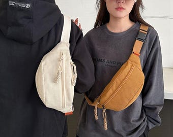 May include: Two corduroy fanny packs in cream and tan. The bags have a zipped main compartment and an adjustable strap with a buckle. The cream bag is worn over a black hoodie, and the tan bag is worn over a grey sweatshirt.