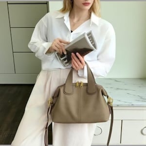 May include: A taupe leather handbag with gold-toned hardware, including zips and clasps. The bag has two top handles and a detachable shoulder strap. The bag is a neutral colour and has a structured shape.
