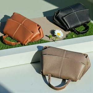 May include: Three leather handbags in shades of brown, black, and taupe. Each bag has a structured design with visible stitching and a small handle. The bags are displayed on a white surface with a patch of green grass and a daisy.