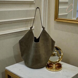 Peut inclure: Un grand sac fourre-tout en daim vert olive avec une poign&eacute;e nou&eacute;e. Le sac est ouvert en haut et a un fond arrondi. Un miroir rond dor&eacute; et des lunettes de soleil reposent sur une surface en marbre blanc.