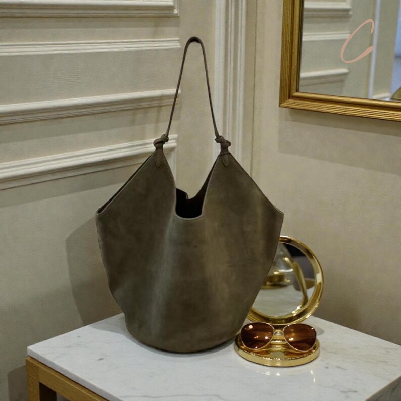 Peut inclure: Un grand sac fourre-tout en daim vert olive avec une poign&eacute;e nou&eacute;e. Le sac est ouvert en haut et a un fond arrondi. Un miroir rond dor&eacute; et des lunettes de soleil reposent sur une surface en marbre blanc.