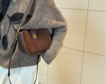 May include: A brown suede shoulder bag with a curved shape and a flap closure. The bag has a long, adjustable strap and gold-coloured hardware. The bag is worn over a brown sweater.
