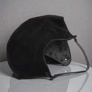 Peut inclure: Un sac &agrave; main en cuir noir avec un design incurv&eacute; unique. Le sac est dot&eacute; d'une seule sangle et d'un int&eacute;rieur textur&eacute;. L'ext&eacute;rieur a une finition mate. Le sac est pos&eacute; sur une surface blanche.