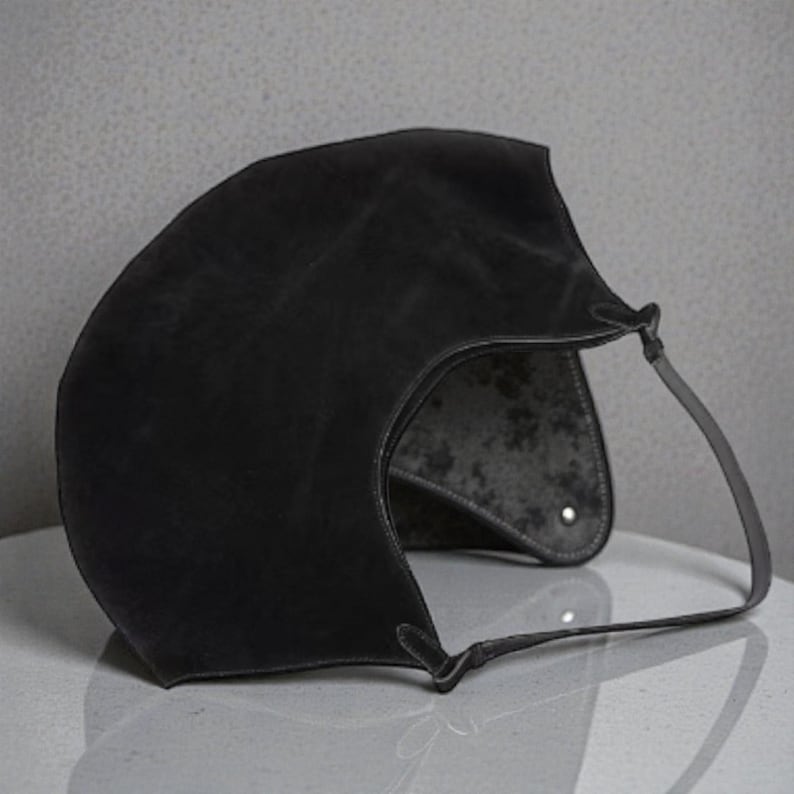 Peut inclure: Un sac &agrave; main en cuir noir avec un design incurv&eacute; unique. Le sac est dot&eacute; d'une seule sangle et d'un int&eacute;rieur textur&eacute;. L'ext&eacute;rieur a une finition mate. Le sac est pos&eacute; sur une surface blanche.