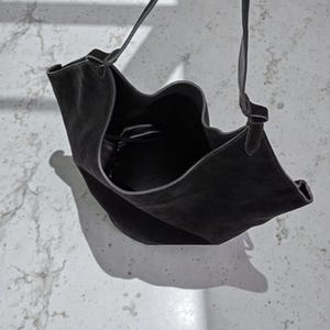 Peut inclure: Sac &agrave; bandouli&egrave;re en daim noir avec une longue sangle. Le sac a un design ouvert unique avec un bord sup&eacute;rieur incurv&eacute;. L'int&eacute;rieur est visible et le sac projette une ombre sur la surface en dessous.
