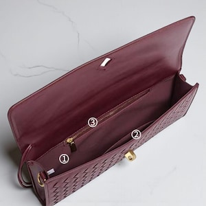 May include: Burgundy leather clutch bag with a woven pattern on the front. Inside, there's a zipped pocket, a slip pocket, and the main compartment. Gold-tone hardware, including the clasp and zip, adds detail.