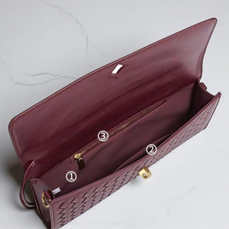 May include: Burgundy leather clutch bag with a woven pattern on the front. Inside, there's a zipped pocket, a slip pocket, and the main compartment. Gold-tone hardware, including the clasp and zip, adds detail.