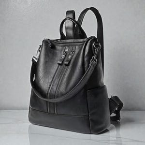 May include: Black leather backpack with multiple zipped compartments and a top handle. The bag features adjustable shoulder straps and a side pocket. The design includes a front pocket with two vertical zips and a crossbody strap.