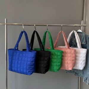 May include: Five quilted tote bags in blue, black, green, pink, and white hang on a metal rack. Each bag has a rectangular shape with a quilted pattern and matching handles. A denim jacket hangs on the right side of the rack.