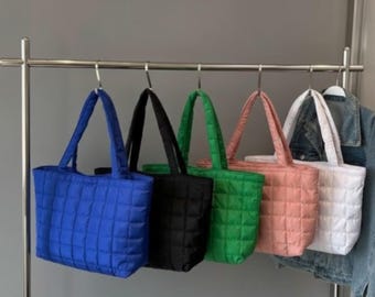 Könnte beinhalten: Fünf gesteppte Tragetaschen in Blau, Schwarz, Grün, Rosa und Weiß hängen an einer Metallstange. Jede Tasche hat eine rechteckige Form mit einem Steppmuster und passenden Griffen. Auf der rechten Seite hängt eine Jeansjacke.