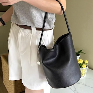 Może przedstawiać: Czarna skórzana torba typu wiadro z długim paskiem i srebrnym łańcuszkiem. Torba ma teksturowaną powierzchnię i zakrzywioną górę. Torba jest noszona na ramieniu.