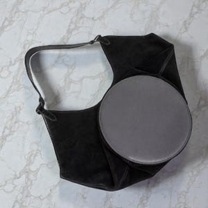Peut inclure: Un sac &agrave; bandouli&egrave;re en daim noir avec un accent circulaire gris. Le sac a une longue sangle et un design unique et pli&eacute;. Le sac est pos&eacute; sur un fond de marbre blanc et gris.