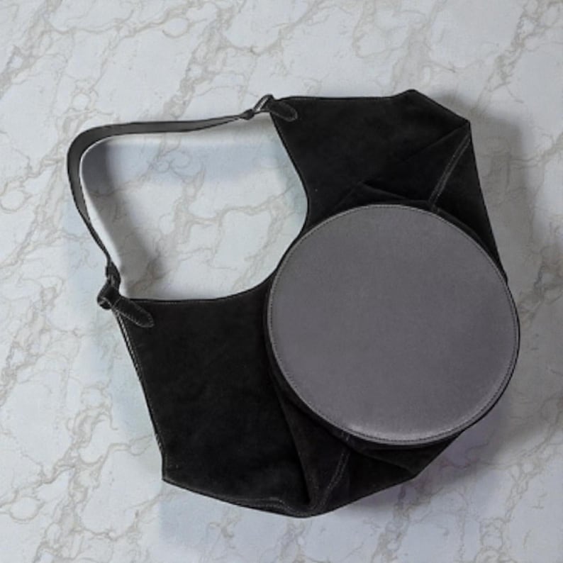 Peut inclure: Un sac &agrave; bandouli&egrave;re en daim noir avec un accent circulaire gris. Le sac a une longue sangle et un design unique et pli&eacute;. Le sac est pos&eacute; sur un fond de marbre blanc et gris.