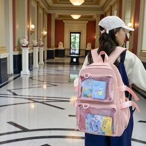 Könnte beinhalten: Ein rosa Rucksack mit transparenten Vordertaschen, in denen farbenfrohe Schreibwaren und Zeichentrickfiguren zu sehen sind. Der Rucksack hat verstellbare Träger und einen oberen Griff. Der Hintergrund ist ein Flur.
