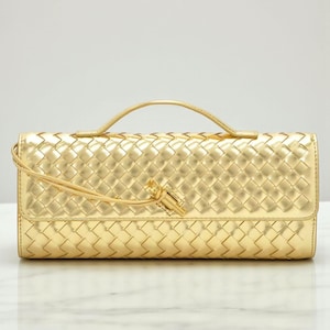 May include: A gold clutch bag with a woven design and a small handle. The bag features a flap closure and a tassel detail. The material is shiny and metallic.