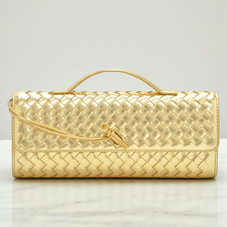 May include: A gold clutch bag with a woven design and a small handle. The bag features a flap closure and a tassel detail. The material is shiny and metallic.