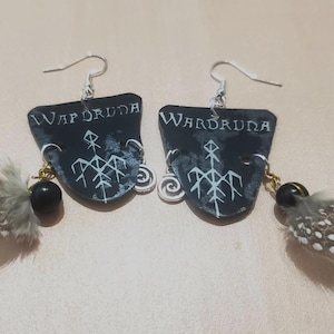 May include: A pair of black shield-shaped earrings with the word "Wardruna" in white script. Each earring features a white graphic, a black bead, and a small feather with brown and white spots. Silver-tone ear hooks.