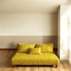 Puede incluir: Un sofá amarillo mostaza con tres cojines a juego. El sofá y los cojines tienen un aspecto texturizado y tejido. La habitación tiene suelos de madera y paredes pintadas en tonos neutros de beige y blanco.