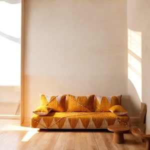 以下が含まれることがあります： 幾何学模様とマッチしたクッションが付いたマスタードイエローのソファ。 ソファはロープロファイルで木製のフレームです。 クッションとソファは、白と黄色の三角形のデザインが特徴です。 ソファの前には小さな木製のサイドテーブルがあります。