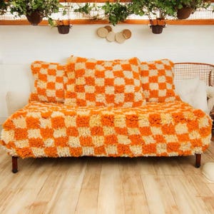 May include: A low-profile sofa with a vibrant orange and cream checkerboard pattern. The sofa has three matching throw pillows and sits on dark wooden legs. The background includes hanging plants and decorative wall accents.