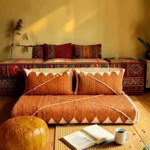 May include: A cozy living room setup featuring a low, orange woven sofa with two matching pillows. The sofa is adorned with white zigzag trim. Additional patterned cushions and a matching ottoman complete the scene, alongside an open book and a coffee mug.