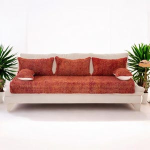 May include: A cream sofa with a rust-coloured, textured cover and matching cushions. The sofa is low-slung and is positioned between potted green plants, creating a comfortable and welcoming living area.