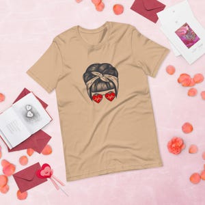 May include: Tan t-shirt featuring a graphic of a woman with her hair up in a bun, wearing heart-shaped sunglasses with a heartbeat design. The shirt is surrounded by pink rose petals and stationery.