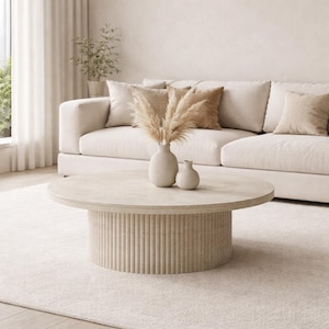 May include: A circular, pale beige coffee table with a ribbed base and a smooth top. Two small, off-white vases hold dried pampas grass. A beige sofa with matching cushions is in the background, creating a neutral, modern living room setting.