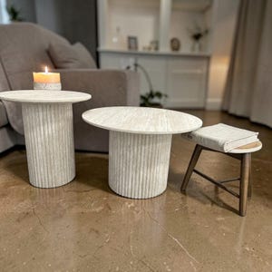 Op de afbeelding: Twee cilindrische bijzettafels met een lichtbeige, getextureerde, gecanneleerde stenen afwerking en ronde bladen. Daarnaast staat een kleine houten kruk met drie schuine poten en een lichtgekleurde stoffen zitting met een boek erop.